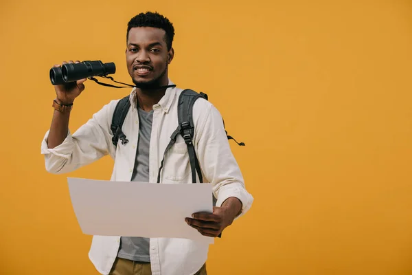 Heureux homme afro-américain tenant des jumelles et du papier blanc isolé sur jaune — Photo de stock
