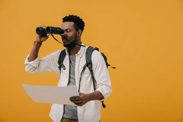 Hombre afroamericano rizado mirando a través de binoculares y sosteniendo papel en blanco aislado en naranja - foto de stock
