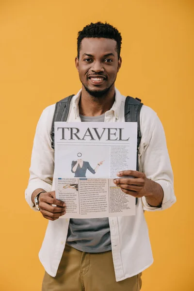 Rizado africano americano hombre de pie con mochila y la celebración de viaje periódico aislado en naranja - foto de stock