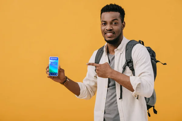Homem americano africano alegre apontando com o dedo para o smartphone com aplicativo de reserva na tela isolado em laranja — Fotografia de Stock