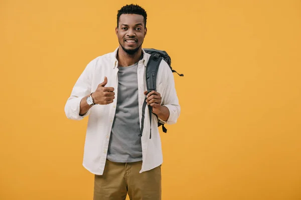 Hombre afroamericano feliz mostrando el pulgar hacia arriba y tocando la mochila aislada en naranja - foto de stock