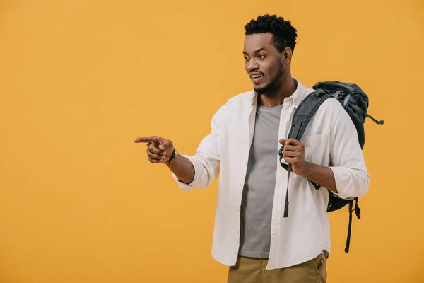 Encaracolado afro-americano apontando com o dedo enquanto em pé com mochila isolada em laranja — Fotografia de Stock