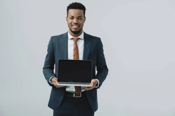 Homme afro-américain joyeux tenant ordinateur portable avec écran blanc isolé sur gris — Photo de stock
