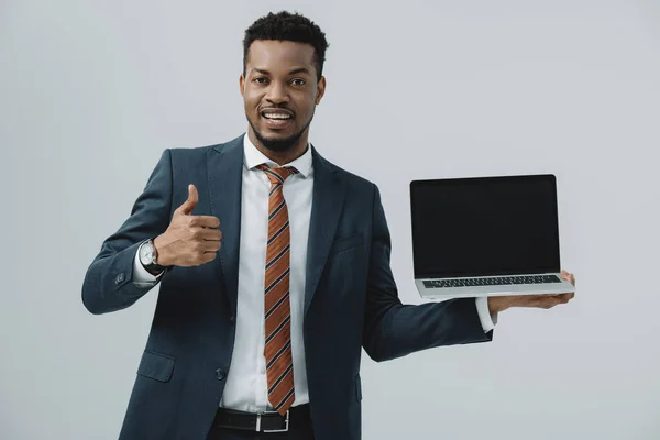 Homme afro-américain joyeux tenant ordinateur portable avec écran blanc et montrant pouce vers le haut isolé sur gris — Photo de stock