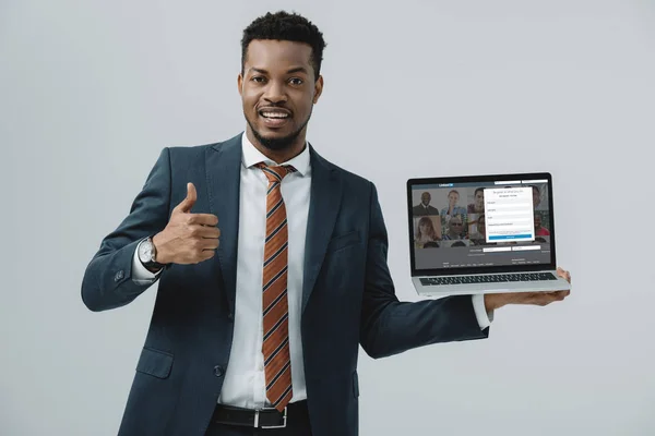 KYIV, UKRAINE - JUNHO 27, 2019: homem americano africano alegre segurando laptop com site linkedin na tela e mostrando o polegar isolado no cinza — Fotografia de Stock