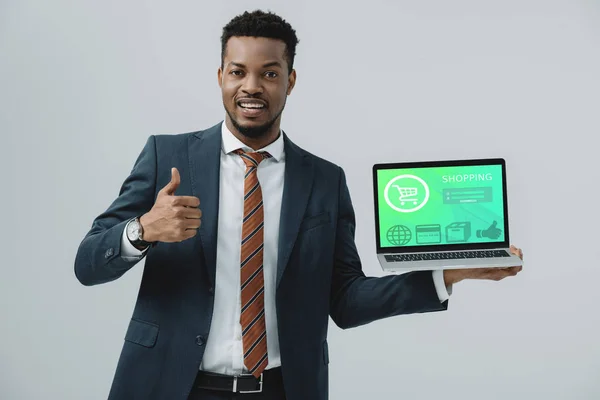 Homme afro-américain joyeux tenant ordinateur portable avec site de shopping à l'écran et montrant pouce vers le haut isolé sur gris — Photo de stock
