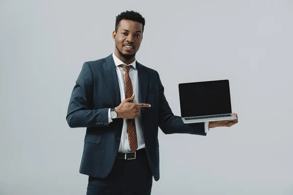 Heureux homme afro-américain pointant avec le doigt à l'ordinateur portable avec écran blanc isolé sur gris — Photo de stock