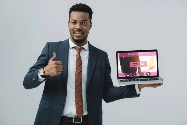 Alegre afroamericano hombre sosteniendo portátil con entradas en línea sitio web en la pantalla y mostrando el pulgar hacia arriba aislado en gris - foto de stock