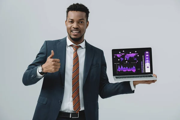 Fröhliche afrikanisch-amerikanische Mann hält Laptop mit Diagrammen und Diagrammen auf dem Bildschirm und zeigt Daumen nach oben isoliert auf grau — Stockfoto