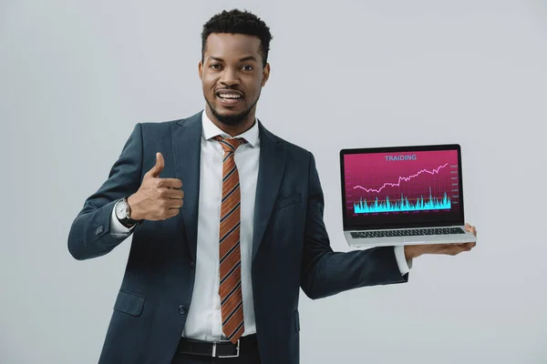 Homme afro-américain joyeux tenant ordinateur portable avec trading analyser à l'écran et montrant pouce vers le haut isolé sur gris — Photo de stock