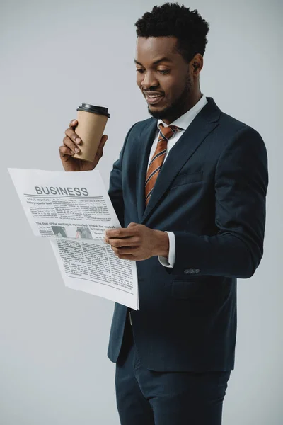 Alegre afroamericano hombre leyendo periódico de negocios y sosteniendo taza de papel aislado en gris - foto de stock