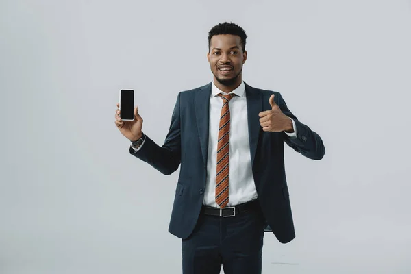 Hombre de negocios afroamericano feliz mostrando el pulgar hacia arriba y sosteniendo el teléfono inteligente con pantalla en blanco aislado en gris - foto de stock