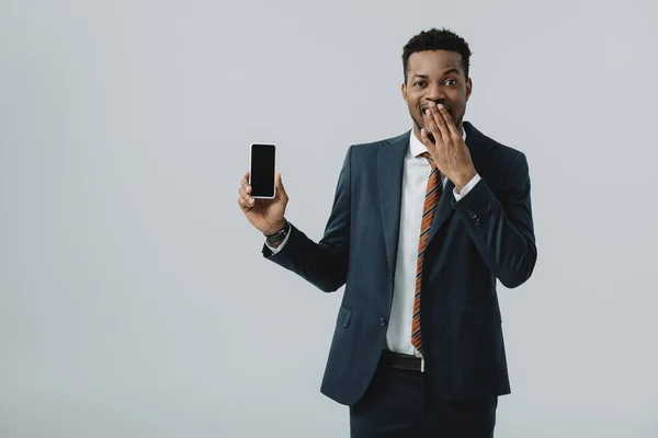 Афроамериканець бізнесмен покриття обличчя і тримає смартфон з порожнім екраном ізольований на сірий — стокове фото