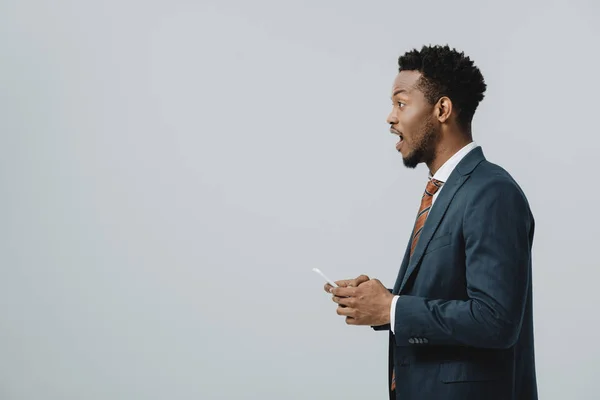 Vista lateral del hombre afroamericano sorprendido utilizando un teléfono inteligente aislado en gris - foto de stock