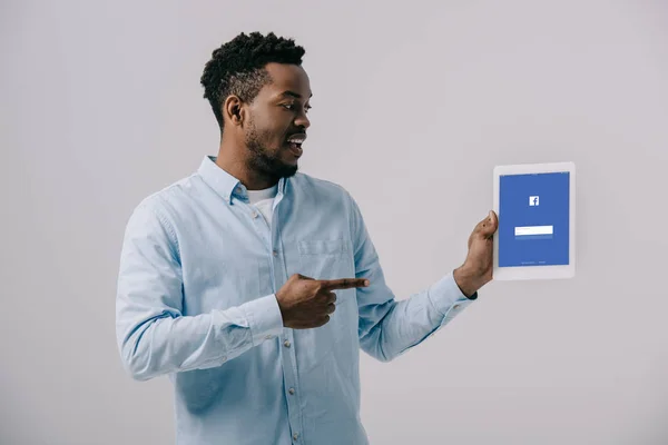 Homem americano africano feliz apontando com o dedo para tablet digital com facebook app na tela isolado em cinza — Fotografia de Stock