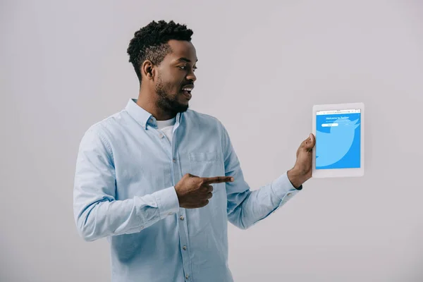 Heureux homme afro-américain pointant du doigt la tablette numérique avec application twitter à l'écran isolé sur gris — Photo de stock