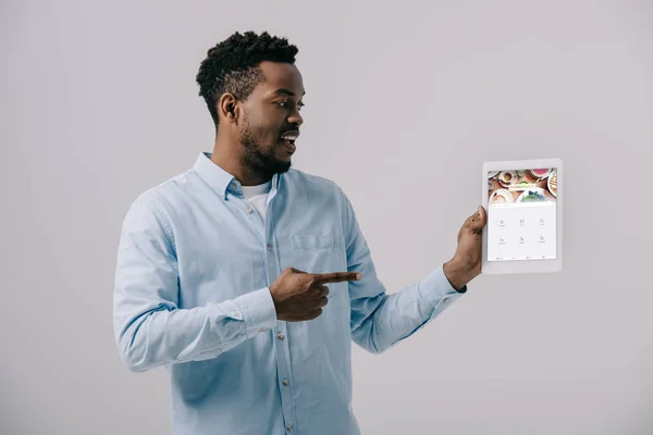 Hombre afroamericano feliz apuntando con el dedo a la tableta digital con la aplicación cuadrada en la pantalla aislada en gris - foto de stock