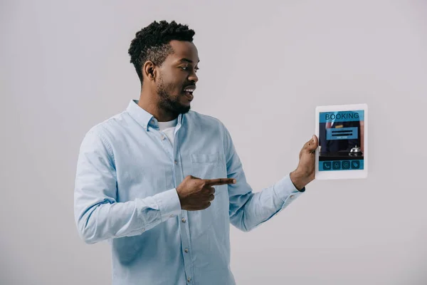Glücklich afrikanisch-amerikanischer Mann zeigt mit dem Finger auf digitales Tablet mit Buchungs-App auf dem Bildschirm isoliert auf grau — Stockfoto