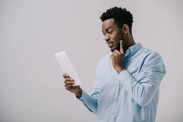 Bell'uomo afro-americano che tocca il viso guardando il tablet digitale isolato sul grigio — Foto stock