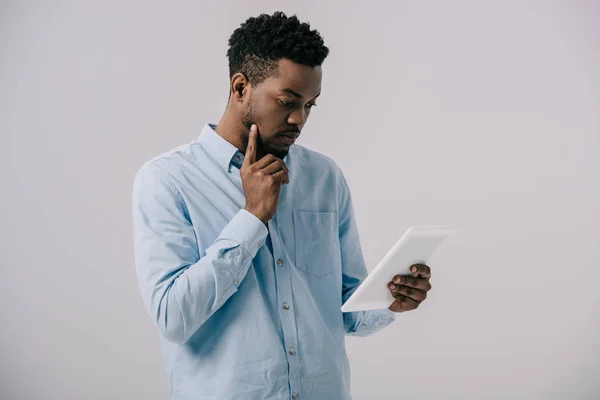 Hombre afroamericano rizado tocando la cara mientras mira la tableta digital aislada en gris - foto de stock