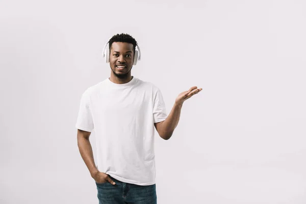 Homem americano africano feliz ouvindo música em fones de ouvido e de pé com a mão no bolso isolado no branco — Fotografia de Stock