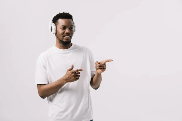 Heureux homme afro-américain écouter de la musique dans les écouteurs et pointant avec les doigts isolés sur blanc — Photo de stock