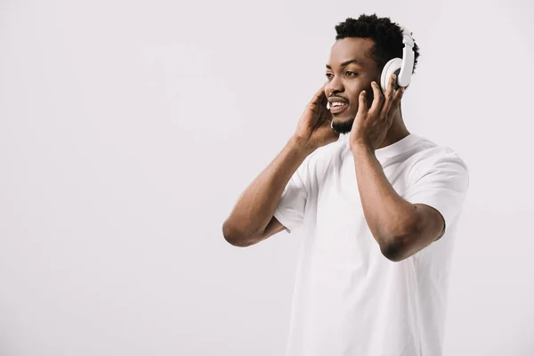 Alegre afroamericano hombre sonriendo y tocando auriculares aislados en blanco - foto de stock