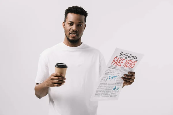 Hombre afroamericano guapo sosteniendo taza de papel y periódico con letras de noticias falsas aisladas en blanco - foto de stock