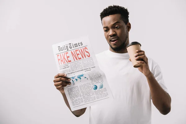 Hombre afroamericano sorprendido sosteniendo taza de papel y leyendo periódico con letras de noticias falsas aisladas en blanco - foto de stock