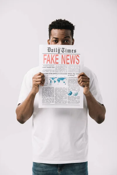 Кудрявый африканский американец, прикрывающий лицо, держа газету с фальшивыми новостями, изолированными на белом — стоковое фото