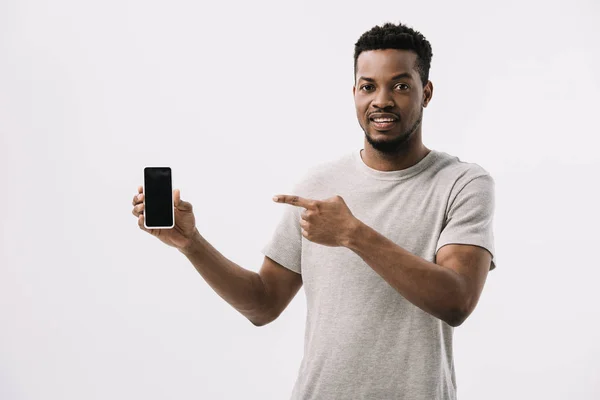 Hombre afroamericano feliz apuntando con el dedo al teléfono inteligente con pantalla en blanco aislado en blanco - foto de stock