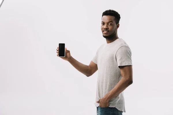 Красивий афроамериканський чоловік стоїть з рукою в кишені і тримає смартфон з порожнім екраном на білому — стокове фото