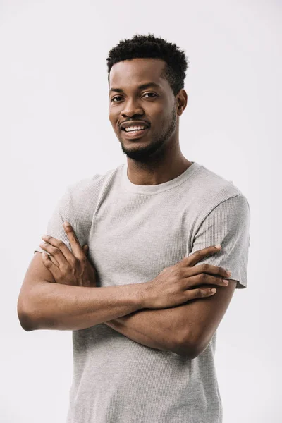 Hombre afroamericano sonriente mirando a la cámara y de pie con los brazos cruzados aislados en blanco - foto de stock
