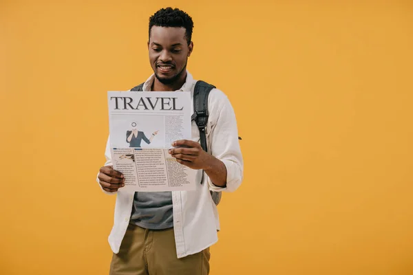 Happy african american man standing with backpack and reading travel newspaper isolated on yellow — Stock Photo