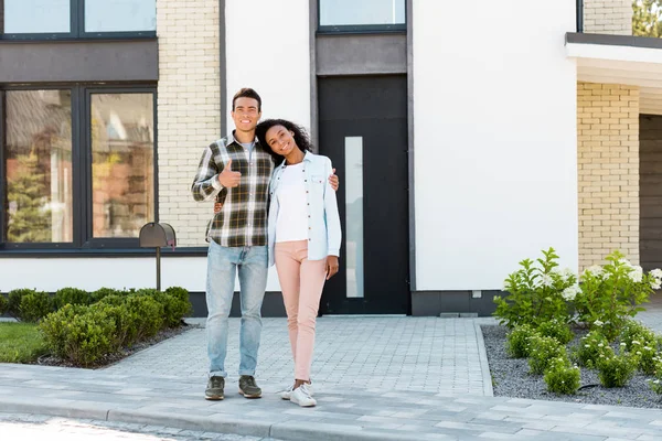 Full length view of african american husband and wife standing near new house and looking at camera while man showing thumb up — Stock Photo