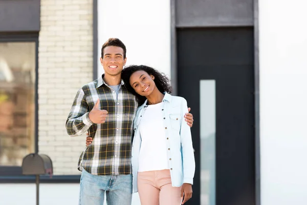 Африканский американский муж и жена стоят рядом с новым домом и смотрят в камеру, в то время как мужчина показывает большой палец вверх — стоковое фото