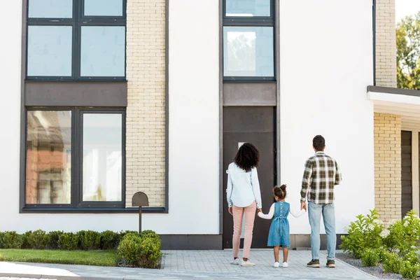 Назад вид африканской американской семьи, стоящей рядом с новым домом, в то время как родители держатся за руки с ребенком — стоковое фото