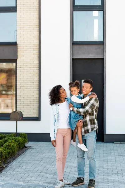 Vista completa de la familia afroamericana de pie cerca de la nueva casa mientras el padre sostiene al niño y la madre mirando al niño - foto de stock