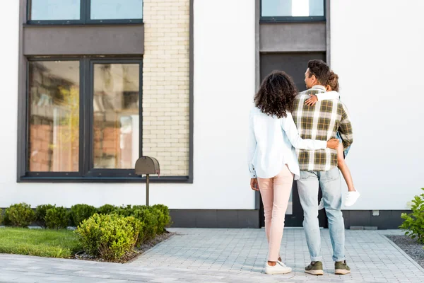 Ansicht der afrikanisch-amerikanischen Familie in voller Länge, die in der Nähe des neuen Hauses steht, während der Vater Kind und Mutter umarmt — Stockfoto