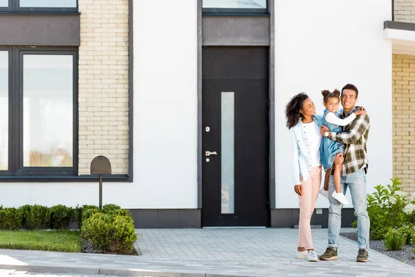 Повнометражний вид на афроамериканську сім'ю, що стоїть біля нового будинку, а батько тримає дитину і дивиться на камеру — стокове фото