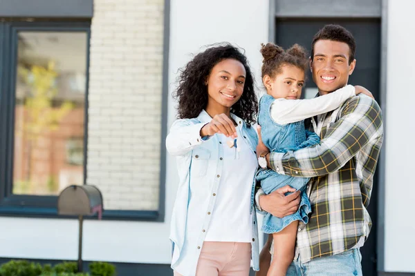 Family standing near new house and looking at camera while father holding kid and mother showing key to camera — Stock Photo