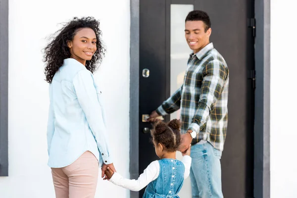 Famille debout près de nouvelle maison tandis que le père tenant la main avec enfant et mère regardant caméra — Photo de stock