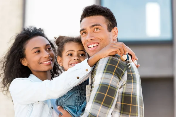 Семья обнимает и смотрит в сторону в то время как африканская американская женщина держит ключ и мужчина держит дочь — стоковое фото