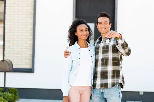 Couple hugging while standing near new house and african american husband presenting key to camera — Stock Photo