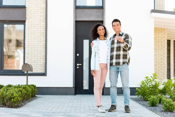 Full length view of couple hugging while standing near new house and husband presenting key to camera — Stock Photo