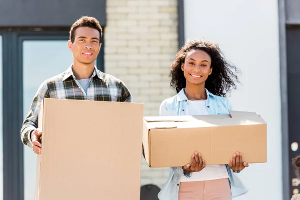 Cople standing near house, looking at camera and holding boxes — Stock Photo