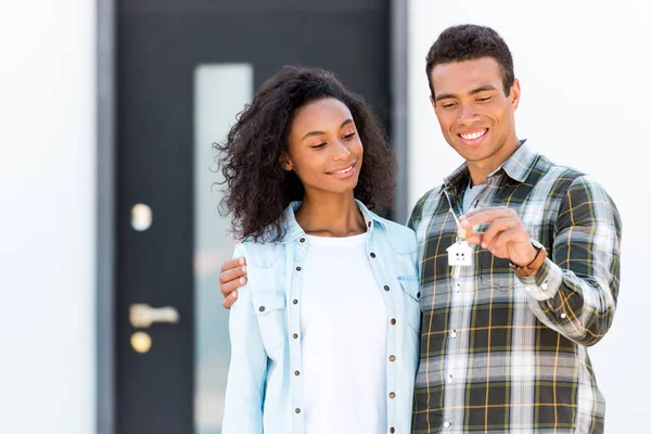 Couple hugging while standing near new house and man holding key to camera — Stock Photo