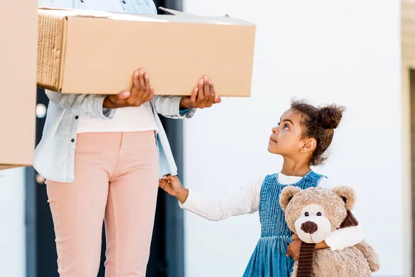 Cropped view of woman holding box while kid standing near mother, holding teddy bear and looking at mom — Stock Photo