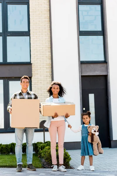 Full length view of african american family standing near house and looking at camera while parent holding boxes and kid holding teddy bear — Stock Photo