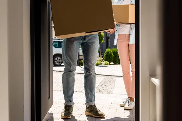 Cropped view of man and woman holding boxes while walking into house — Stock Photo